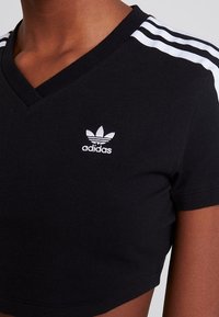 Černé zkrácené tričko vyrobené z bavlny, s V-čelní výstřihem a bílým logem Adidas se třemi bílými pruhy na ramenou.