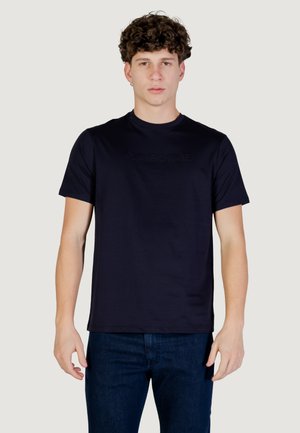 Giovane uomo con capelli ricci che indossa una t-shirt Armani Exchange a maniche corte blu scuro e jeans blu scuro su uno sfondo chiaro.