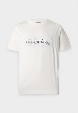 Homme portant un t-shirt blanc Emporio Armani, un pantalon marron à jambes larges et des chaussures noires, debout devant un fond clair uni.