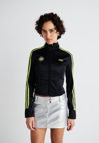 Jachetă neagră cu fermoar, având dungi de un verde aprins pe mâneci, cu un patch cu logo. Asortată cu o fustă mini texturată de culoare argintie.