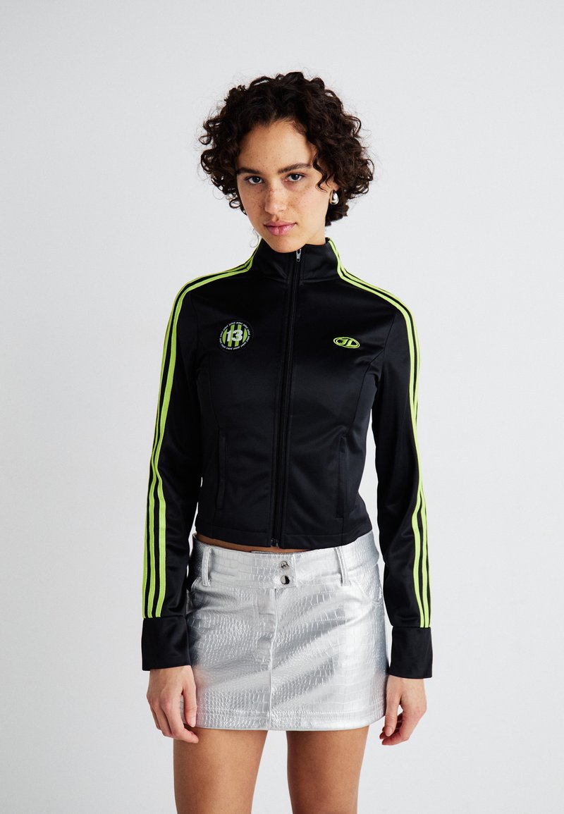 Jachetă neagră cu fermoar, având dungi de un verde aprins pe mâneci, cu un patch cu logo. Asortată cu o fustă mini texturată de culoare argintie.