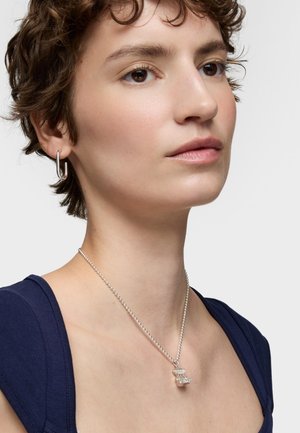 Collana in argento con un pendente attorcigliato. Il modello ha i capelli corti e ricci e indossa una maglietta blu navy. Orecchino ad anello in un orecchio.