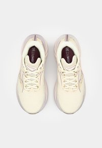 Lys beige treningssko med en teksturert mesh-overdel, flate lisser og rosa detaljer. Har en polstret såle og ventilasjonsåpninger.