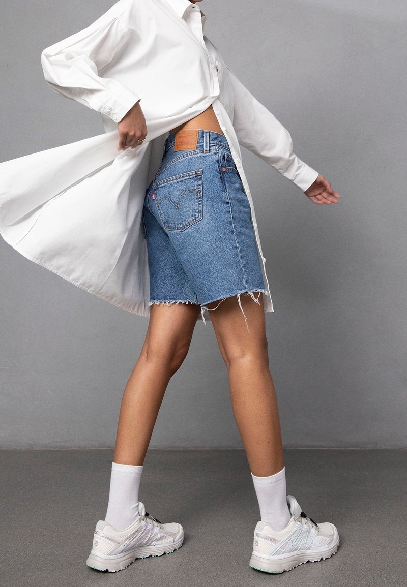 Lichtblauwe denimshorts met rafelige zoom, gecombineerd met een wit shirt met lange mouwen. Witte sneakers met grijze accenten en witte sokken.