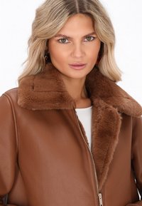 Kvinde med blond hår iført en brun læderjakke med en tyk, fluffy krave, der ser lige frem mod en hvid baggrund.