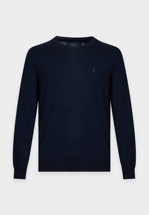 Homme portant une chemise bleu marine à manches longues, pantalon blanc et chaussures noires, debout avec une main dans la poche devant un fond blanc uni.
