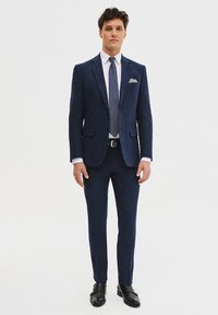 Granatowy garnitur z teksturowanym wykończeniem, biała koszula, krawat w kropki i poszetka, w zestawieniu z czarnymi butami. Slim-fit design z dopasowanymi spodniami.