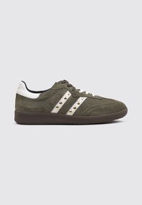 Zapatillas de ante verde con acentos en cuero blanco, que presentan tres rayas blancas con tachuelas doradas, punta redonda y suela de goma texturizada marrón.