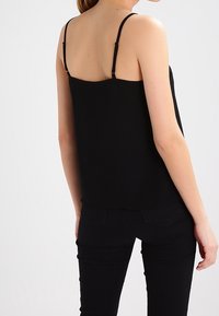 Kvinna iklädd en svart ärmlös topp med tunna remmar och svarta åtsittande byxor, sedd bakifrån mot en enfärgad ljus bakgrund.