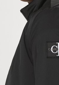 Doudoune noire avec un col montant, présentant une texture matelassée, une fermeture éclair et un patch de marque bien visible sur la poitrine.