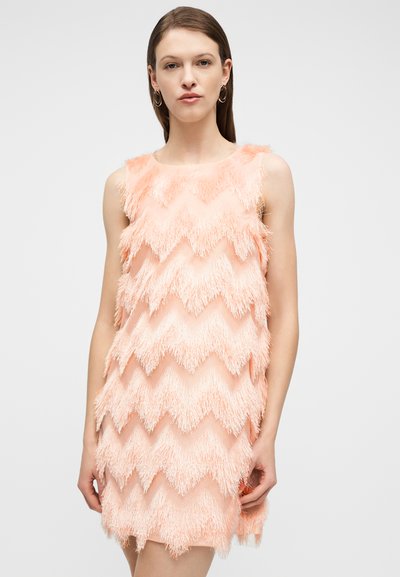 Γυναίκα με ίσια καστανά μαλλιά που φοράει αμάνικο ροδακινί φόρεμα με υφασμάτινο μοτίβο ζιγκ-ζαγκ, στέκεται μπροστά από απλό φωτεινό φόντο.