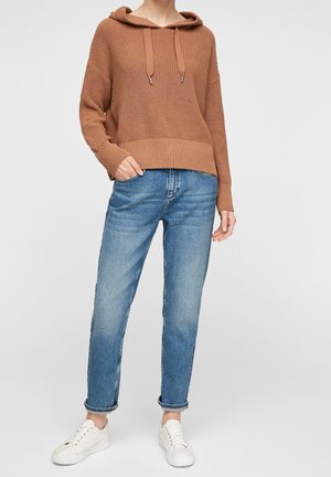 Personne portant un sweat à capuche en maille marron, un jean bleu avec revers et des baskets blanches, debout devant un fond clair uni.