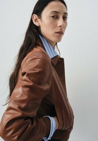 Veste en cuir marron au design structuré, ornée d'un col de chemise rayé bleu et blanc et de poignets côtelés, sur un fond neutre.