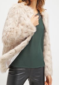 Faux furjakke i blød beige, med lange ærmer, en afslappet pasform og struktureret overflade. Sammen med en mørkegrøn top og strømlinede sorte bukser.