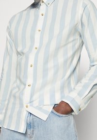 Camicia button-up a righe azzurro chiaro e bianco con bottoni dorati, colletto classico e vestibilità leggermente attillata. Il tessuto in cotone offre una texture morbida.