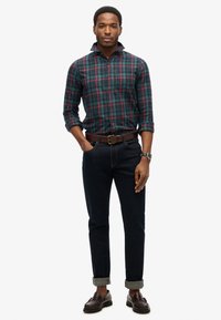 Groen en marineblauw ruitjesoverhemd met knopen, donkere jeans opgerold bij de boorden, bruine leren riem en bruine loafers. Horloge om de linkerpols.