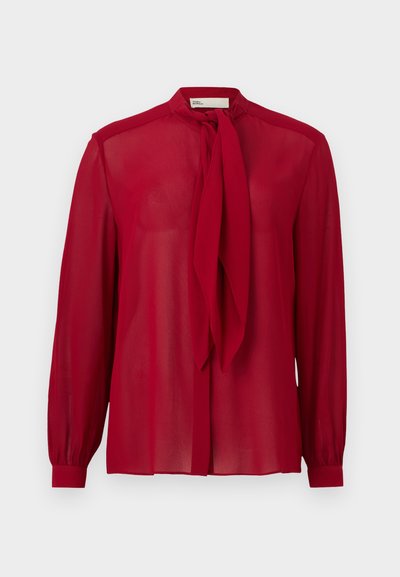 Blouse rouge transparente à manches longues avec un nœud à nouer au col et des poignets à boutons, présentée sur un fond clair uni.
