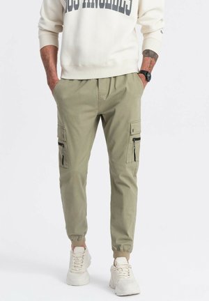 Pantalones cargo en verde oliva, con puños elásticos y una cinturilla ajustable, con cuatro bolsillos, incluidos dos con cremallera. Se lleva con un sudadera color crema.