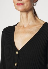Černý žebrovaný cardigan s hlubokým výstřihem do V, který má vpředu dva kulaté knoflíky. Tkanina vypadá na dotek měkce a má strukturovaný povrch.