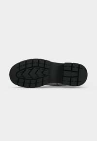 Semelle d'une botte noire avec des crampons profonds, présentant des motifs en chevrons et rectangulaires pour une bonne adhérence sur un fond uni.