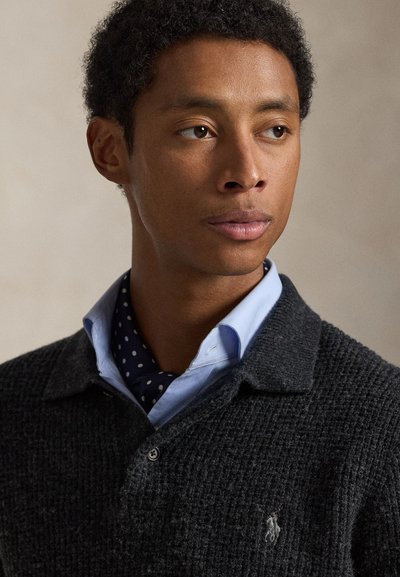 Dark textured knit sweater with a collar, worn over a light blue shirt and a navy polka dot scarf. Features a small logo on the sweater.