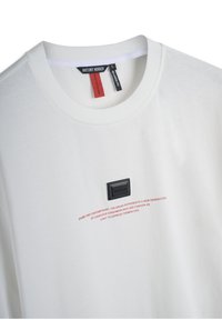 Camiseta blanca de algodón con cuello redondo, que presenta una etiqueta rectangular negra en el pecho y texto rojo impreso debajo de la etiqueta.