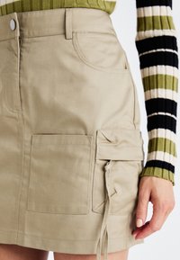 Béžová cargo sukně s přiléhavým střihem, vybavená předními kapsami a nastavitelnými bočními šňůrkami, doplněná zeleným proužkovaným tričkem s dlouhým rukávem.