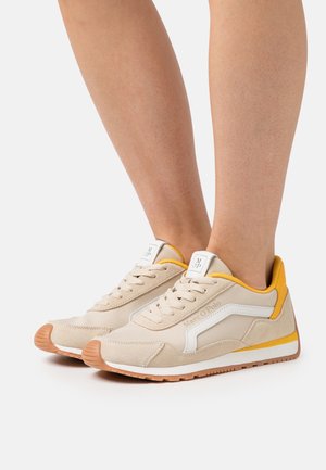 Béžové a bílé tenisky se žlutými akcenty a gumovými podrážkami, nošené na bosých nohou, s logem "Marc O'Polo" na boku.