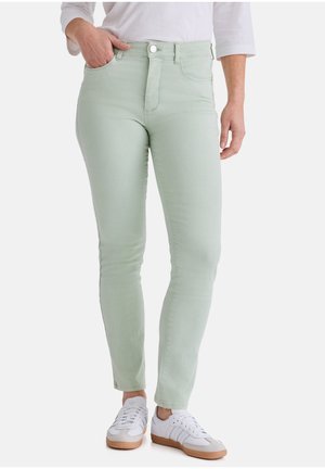 Person trägt hellgrüne Slim-Fit-Jeans mit Taschen vorne, kombiniert mit weißen Sneakern und einem weißen Oberteil, steht vor einem schlichten Hintergrund.