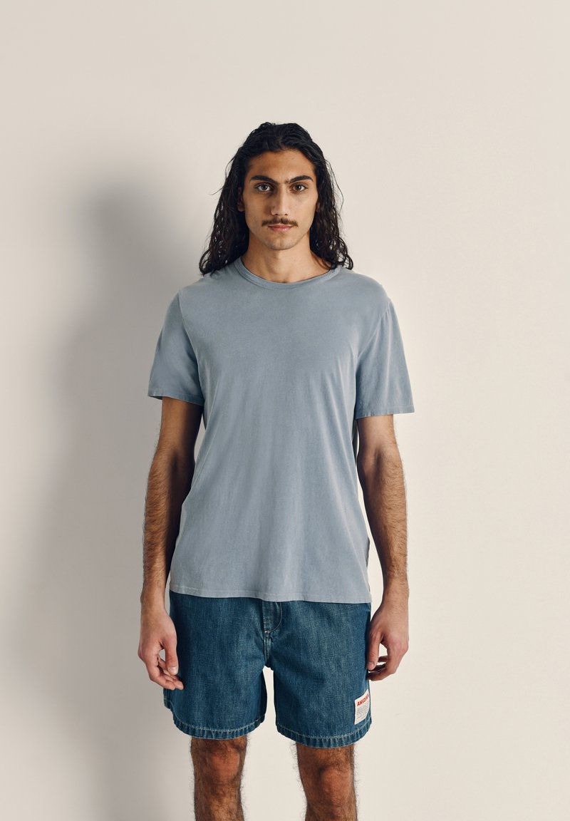 Light blue short-sleeve t-shirt in smooth fabric, paired with dark denim shorts featuring a side label. Simple, casual design.