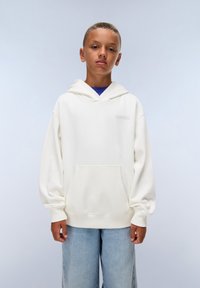 Un sweat à capuche crème avec une poche frontale, des manches longues et un cordon de serrage pour la capuche. Porté sur une chemise bleue et associé à un jean bleu clair.