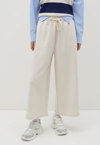 Pantaloni tuta beige a gamba larga con vita regolabile, realizzati in un tessuto morbido. Abbinati a un top a strati blu chiaro e scarpe da ginnastica bianche.