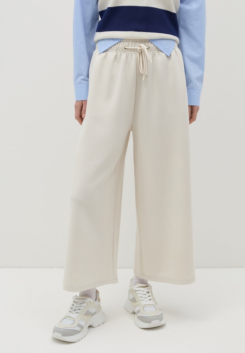 Pantaloni tuta beige a gamba larga con vita regolabile, realizzati in un tessuto morbido. Abbinati a un top a strati blu chiaro e scarpe da ginnastica bianche.