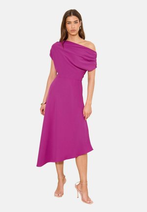 Naine kannab varrukateta, ühe õla paljastavat, asümmeetrilist magenta keskmise pikkusega kleiti, kumeraid kõrge kontsaga sandaale ja kuldseid rõngakujulisi kõrvarõngaid.