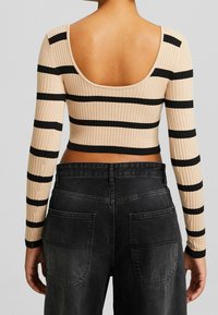Geripptes, kurzes Top in Beige mit horizontalen schwarzen Streifen. Verfügt über einen weiten Rundrücken und lange Ärmel. Kombiniert mit hochgeschnittenen schwarzen Jeans.