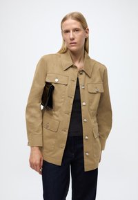 Chaki spalvos medvilninė striukė su keturiomis priekinėmis kišenėmis, metalinėmis spaustukėmis ir tiesia kirpimo linija, rankoje laikantis juodą delninę.