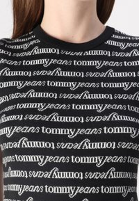 Haut en tricot noir avec un motif de logo blanc "tommy jeans" répété. Encolure ronde structurée et manches courtes. Texture lisse.