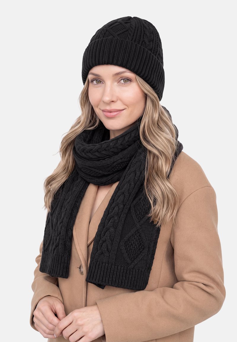Mujer con cabello largo, ondulado y rubio, que lleva un gorro tejido negro y una bufanda a juego, y un abrigo marrón claro, sonriendo ligeramente contra un fondo liso.