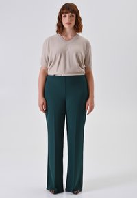 Vrouw met kort golvend kastanjebruin haar, gekleed in een beige V-hals korte mouwen top en donkergroene hoge taille wijde broek, tegen een effen achtergrond.