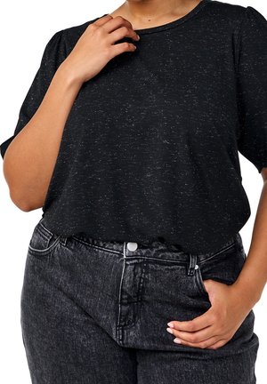 Personne portant un t-shirt noir moucheté rentré dans un jean en denim foncé taille haute, une main près du col et l'autre dans une poche.