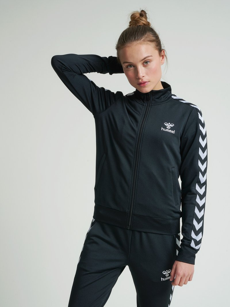 Kvinde med hår i en knold iført en sort sportsdragt med hvide chevronstriber og "hummel"-logo, stående mod en ensfarvet baggrund.