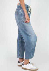 Personne portant un jean en denim bleu bicolore avec des poches plaquées, des baskets blanches avec des accents noirs, et un haut blanc avec un cordon de serrage à la taille.