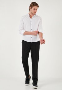 Camisa blanca de lino de manga larga con botones, pantalones negros a medida y zapatillas negras. La camisa tiene un cuello alzado y botones marrones.