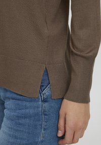 Pull en tricot marron à manches longues, avec des fentes sur les côtés à l'ourlet, associé à un jean en denim bleu, affichant une texture lisse.