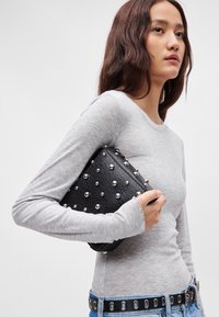 Čierna kožená kabelka na ruku s striebornými klincami, obdĺžnikový tvar, textúrovaný povrch, držaná pri šedej košeli s dlhým rukávom a modrých džínsach.