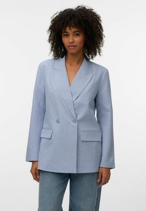 Woman with curly hair wearing a light blue double-breasted blazer and blue jeans, standing against a plain white background.