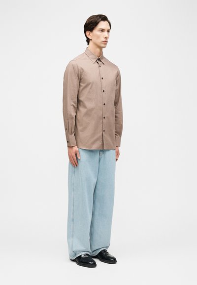 Άνδρας στέκεται με το πρόσωπο προς τα εμπρός φορώντας ένα μπεζ πουκάμισο με κουμπιά, ανοιχτό μπλε τζιν με φαρδιά μπάτζια και μαύρα επίσημα παπούτσια σε λευκό φόντο.