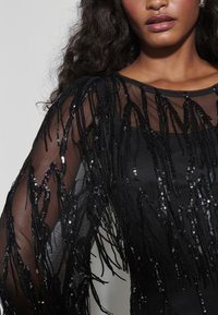 Robe noire en tissu transparent avec des franges longues et pendantes ornées de perles. La texture est lisse avec des sequins réfléchissants partout.
