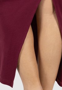 Primer plano de piernas desnudas visibles a través de una abertura lateral en una falda o vestido acanalado de color rojo oscuro.