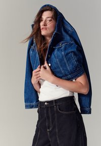 Kék farmerdzseki kapucnival, ezüst gombokkal és zsebekkel, fehér ujjatlan felső és fekete magasderekú nadrág fölött viselve.